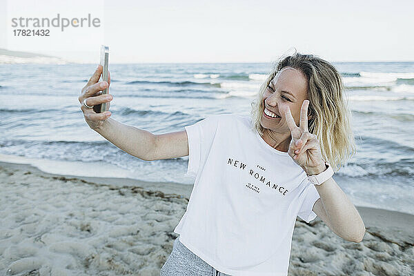 Glückliche Frau gestikuliert ein Friedenszeichen und macht am Strand ein Selfie per Smartphone
