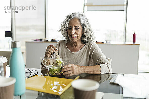 Glückliche Geschäftsfrau mit grauen Haaren  die am Schreibtisch im Büro Salat isst