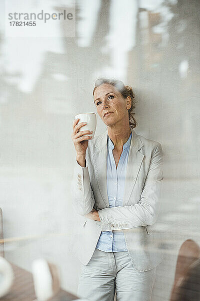 Nachdenkliche Geschäftsfrau hält Kaffeetasse im Café  gesehen durch Glas