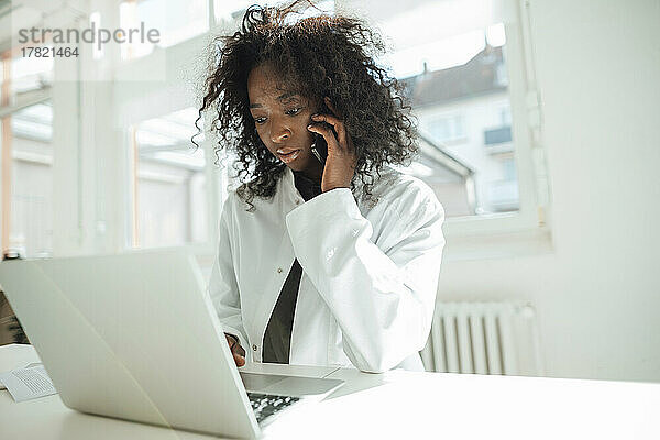 Ärztin spricht mit Smartphone und Laptop am Schreibtisch