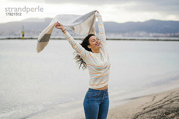 Fröhliche Frau mit erhobenen Armen und Schal am Strand