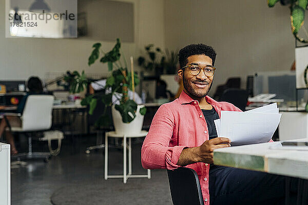 Lächelnder Geschäftsmann mit Dokumenten am Schreibtisch im Büro