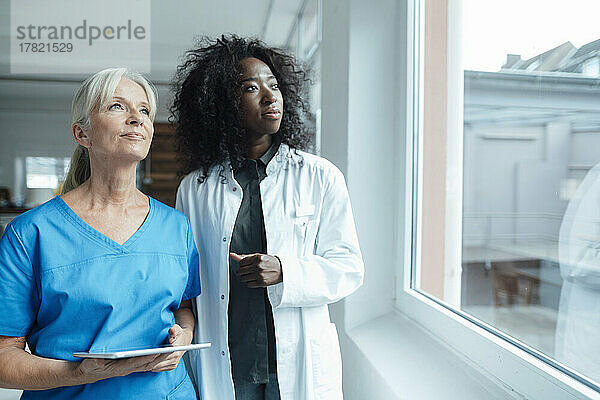 Krankenschwester hält Tablet-PC in der Hand und schaut neben Ärztin durchs Fenster