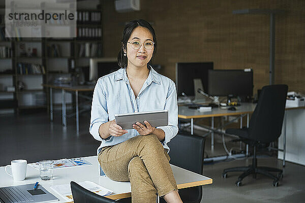 Lächelnde Geschäftsfrau mit Brille sitzt am Schreibtisch am Arbeitsplatz