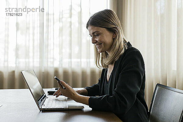 Lächelnde Geschäftsfrau mit Mobiltelefon und Laptop auf dem Tisch