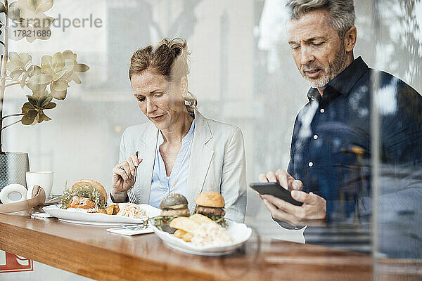 Geschäftsfrau isst zu Mittag und sitzt neben einem Geschäftsmann  der sein Smartphone im Café benutzt und durch Glas gesehen wird