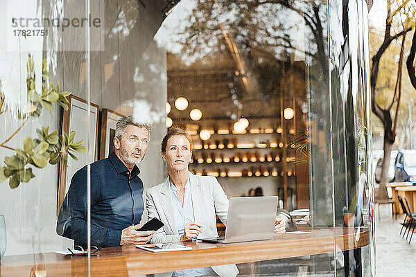 Geschäftsmann hält Mobiltelefon in der Hand und steht Geschäftsfrau im Café gegenüber  die durch das Fenster gesehen wird