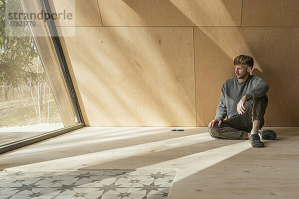 Mann sitzt auf dem Boden in einem Öko-Holzhaus und schaut aus dem Fenster