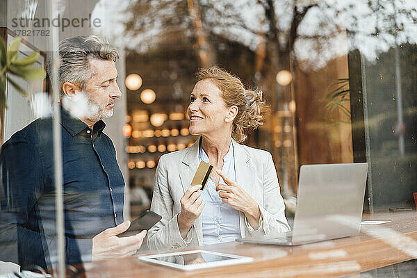 Geschäftsfrau zeigt auf Kreditkarte und diskutiert mit Geschäftsmann im Café  gesehen durch Glas