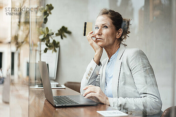 Geschäftsfrau mit Kreditkarte sitzt mit Laptop im Café und sieht durch Glas