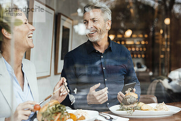 Glückliche Geschäftsfrau mit Geschäftsmann  der sein Mobiltelefon in der Hand hält und im Café zu Mittag isst  gesehen durch Glas