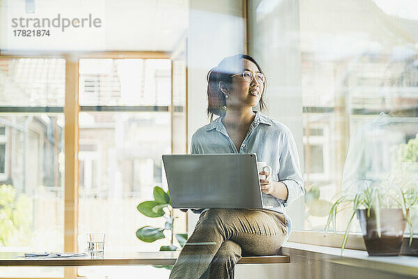 Smiling woman with laptop sitting on desk by window