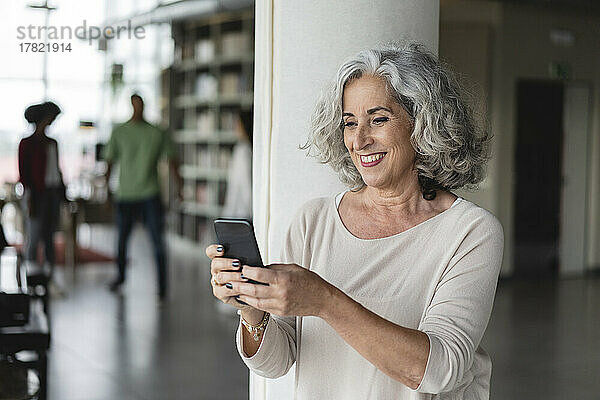 Lächelnde Geschäftsfrau nutzt Smartphone per Säule im Büro