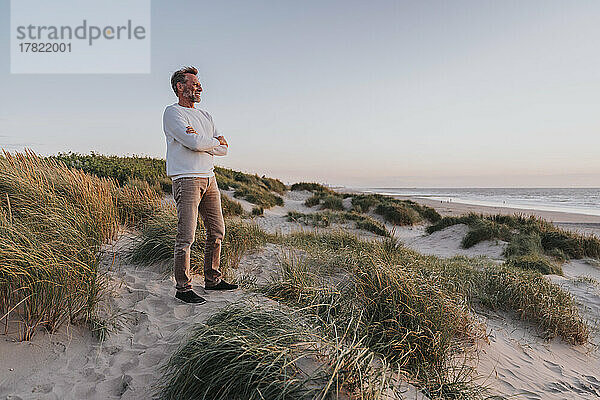 Happy man with arms crossed standing on sand dune at beach