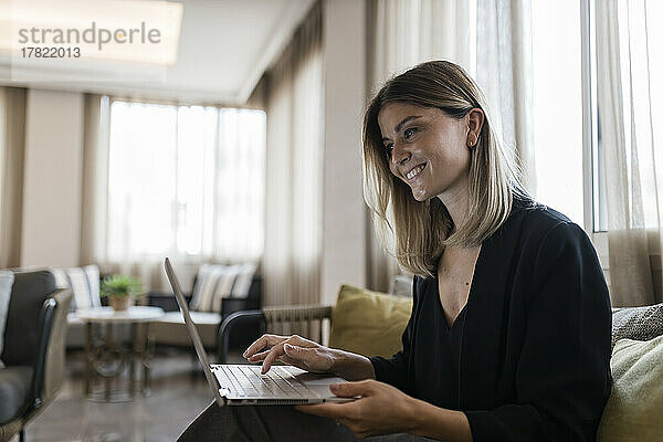 Lächelnde Geschäftsfrau sitzt mit Laptop auf dem Sofa in der Hotellobby