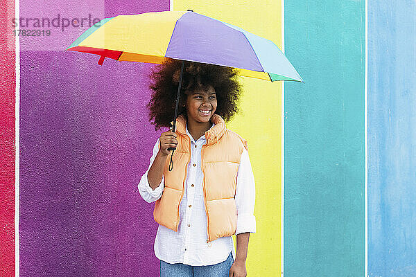 Fröhliches Mädchen steht mit buntem Regenschirm vor einer bunten Wand