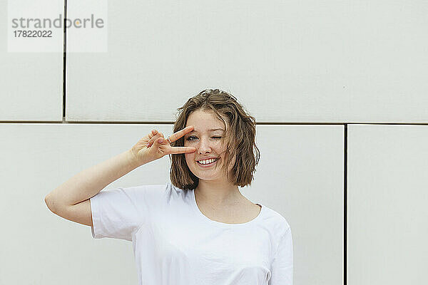 Lächelndes Teenager-Mädchen zwinkert mit den Augen und gestikuliert ein Friedenszeichen  das vor der Wand steht