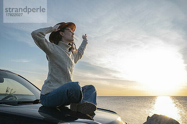 Junge Frau mit Hut sitzt auf der Motorhaube eines Autos