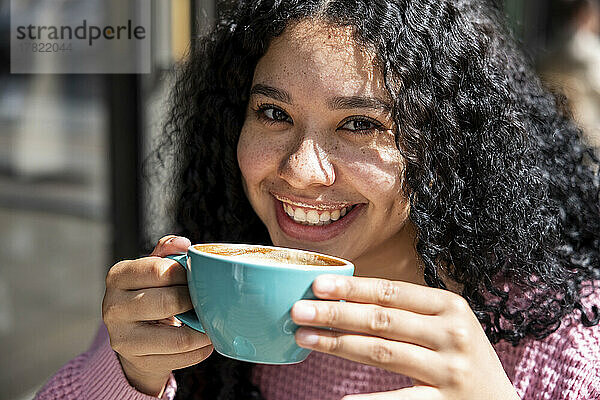 Glückliche junge Frau mit lockigem Haar hält an einem sonnigen Tag eine Kaffeetasse in der Hand