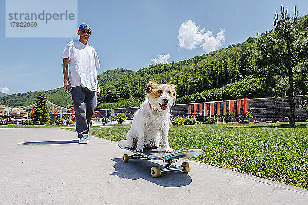 Lächelnder Mann mit Hund sitzt auf Skateboard im Park