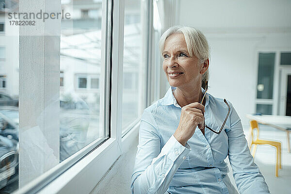 Lächelnde ältere Geschäftsfrau mit Brille und Blick durch das Fenster im Büro