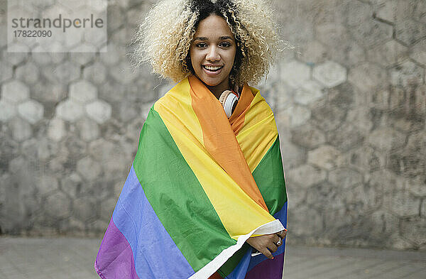 Lächelnde Frau mit Regenbogenfahne bedeckt  die vor der Wand steht