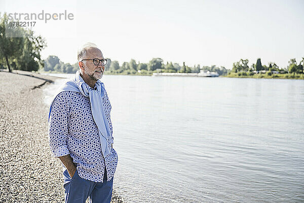 Älterer Mann steht mit den Händen in den Taschen am Flussufer