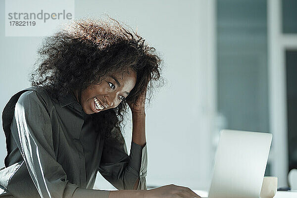 Happy businesswoman with hand in hair using laptop at office