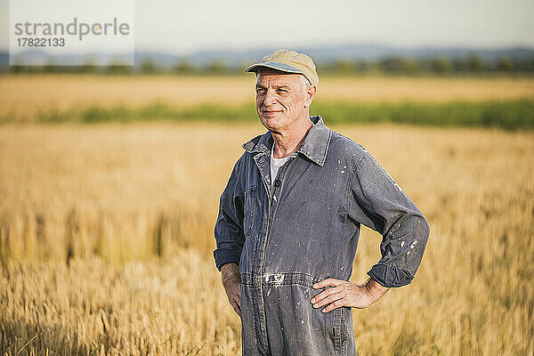 Lächelnder älterer Bauer mit Mütze steht mit in die Seite gestemmten Armen auf dem Bauernhof