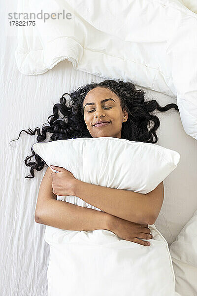 Glückliche Frau mit geschlossenen Augen umarmt Kissen auf dem Bett im Schlafzimmer
