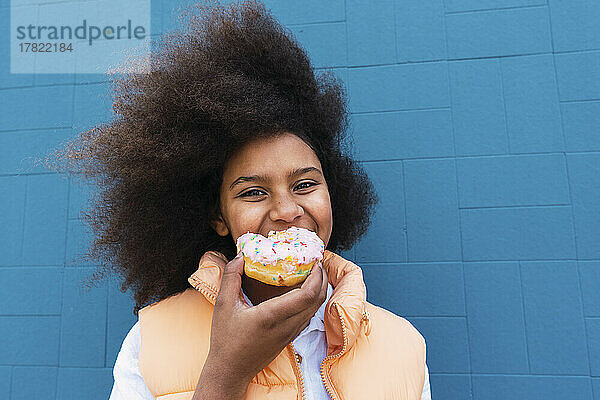 Lächelndes Mädchen isst Donut vor blauer Wand
