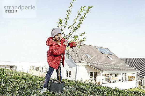 Lächelndes Mädchen umarmt Topfpflanze vor Haus