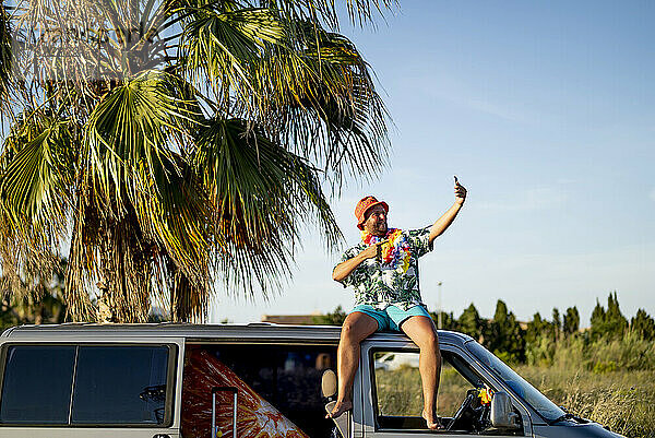 Glücklicher Mann  der auf dem Dach eines Lieferwagens sitzt und ein Selfie mit seinem Mobiltelefon macht