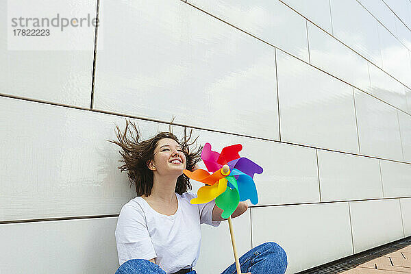Fröhliches Teenager-Mädchen mit buntem Windrad-Spielzeug sitzt vor der Wand