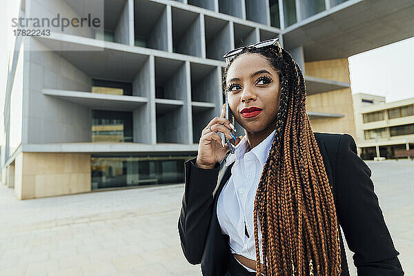 Lächelnde junge Geschäftsfrau mit Smartphone im Gespräch vor Bürogebäude