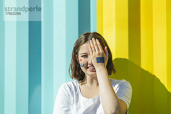 Lächelndes Teenager-Mädchen mit Friedenssymbolfarbe im Gesicht  das das Auge vor einer blauen und gelben Wand bedeckt