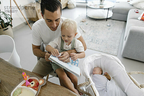 Vater hält Buch und sitzt mit Sohn neben Baby  das zu Hause im Kinderbett schläft