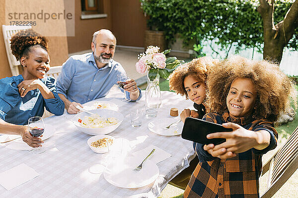 Lächelnder Junge macht ein Selfie mit der Familie per Smartphone am Esstisch im Hinterhof