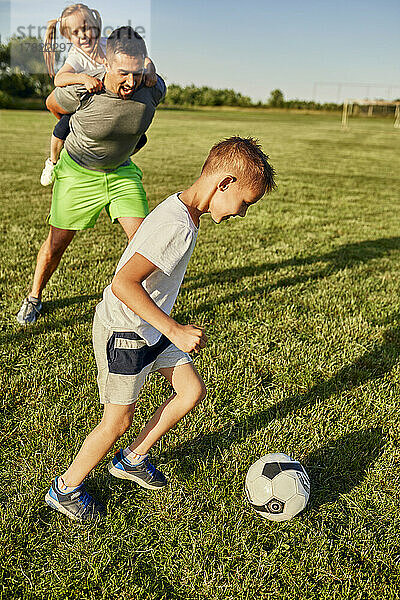 Junge spielt Fußball  während ein Mann einem Mädchen auf dem Sportplatz Huckepackfahrt gibt