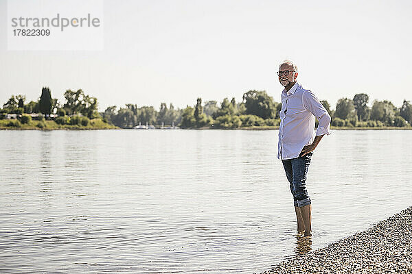 Lächelnder Mann steht mit der Hand auf der Hüfte am Flussufer
