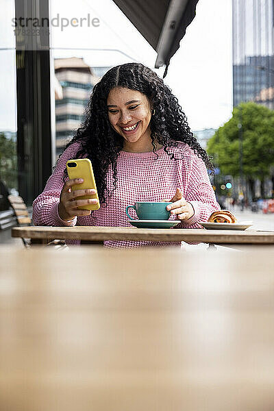 Glückliche Frau  die mit ihrem Mobiltelefon im Internet surft und mit einer Kaffeetasse im Straßencafé sitzt