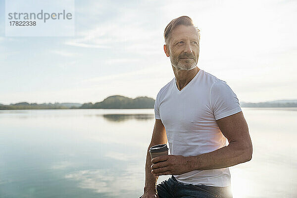 Nachdenklicher reifer Mann sitzt an einem sonnigen Tag mit einer Kaffeetasse am See