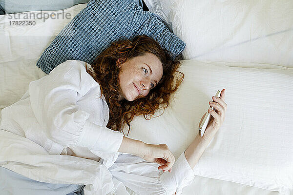 Frau mit braunen Haaren benutzt Smartphone im Bett liegend