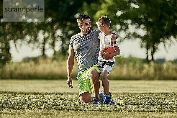 Junge hält Rugbyball und unterhält sich mit Vater  der auf dem Sportplatz kniet