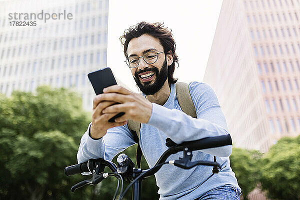 Lächelnder Mann benutzt Mobiltelefon und stützt sich auf Fahrrad