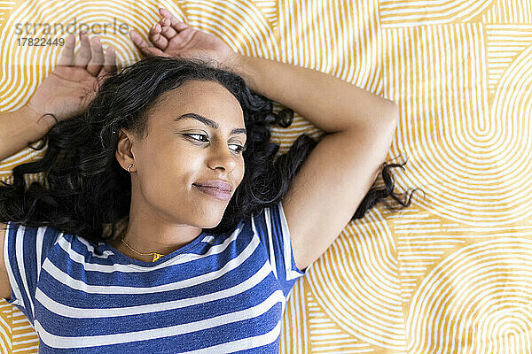 Thoughtful woman with arms raised lying on bed at home
