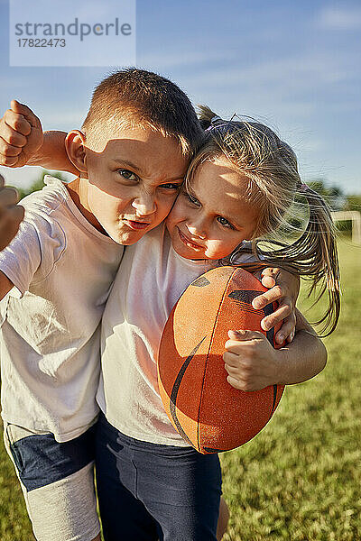 Junge macht Gesicht und steht mit Schwester  die an einem sonnigen Tag Rugbyball hält