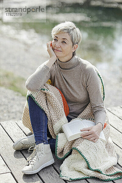 Nachdenkliche Frau sitzt mit Buch und Strickdecke am Pier