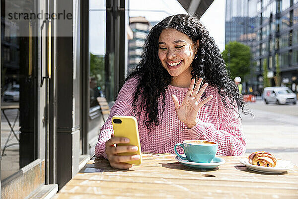 Lächelnde Frau winkt bei einem Videoanruf über ihr Smartphone im Straßencafé mit der Hand