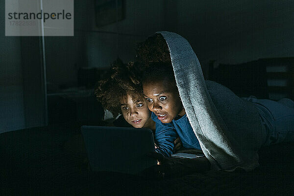 Shocked woman looking at tablet PC with daughter under blanket